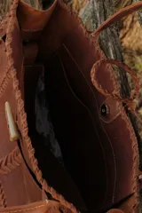 Bolso tipo tote de cuero color marrón con detalles de costuras trenzadas en los bordes y bolsillo externo. Tiene cierre imantado interno y broche con pasador de guampa en el exterior.