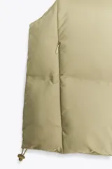 Chaleco blanco acolchado con cuello alto y cierre frontal con cremallera. Tiene bolsillos delanteros con cierre de cremallera oculta y bajo ajustable con cordones elásticos laterales.