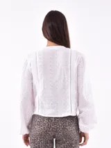 Camisa blanca con bordado calado, cuello redondo, mangas largas abullonadas con puños elásticos y cierre frontal con botones.