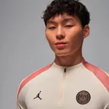 Remera de manga larga color crema con detalles en salmón en los hombros, con logo de Jordan y escudo del Paris Saint-Germain en el pecho y leyenda "PARIS" estampada verticalmente en la manga izquierda.