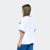 Remera blanca de mujer con cuello y mangas con detalles tricolor (azul, blanco y rojo), logo de Umbro bordado en negro y escudo del Club Nacional de Football estampado.
