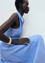 Vestido midi de algodón y lino, color celeste con rayas verticales blancas. Presenta cuello camisero, diseño sin mangas con tirantes anchos, cierre de botones frontales, cinturón con hebilla y falda evasé.