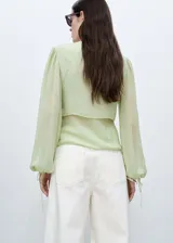 Blusa color verde claro de gasa semitransparente con cuello redondo y lazos en el frente. Tiene mangas largas abullonadas con puños elásticos y fruncidos. Incluye un chaleco corto incorporado. 