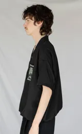 Remera oversize negra de piqué de algodón con bolsillo estampado con una palmera Phoenix Canariensis. Tiene elástico verde con hebilla de ajuste en la parte inferior.