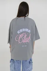 Remera gris de corte oversize con cuello redondo y estampado pequeño en el pecho con la frase "Karma Club" en color lila.