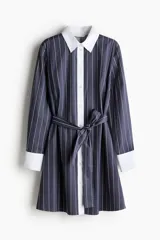 Vestido corto estilo camisero a rayas verticales azul marino y blancas, con cuello y puños blancos. Tiene cierre frontal con botones y lazo para atar en la cintura.