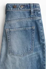 Jeans de tiro alto en denim de algodón rígido, color azul medio con efecto degradado (ombré) que se oscurece hacia el bajo. Corte acampanado (flare) y bajo deshilachado. Presenta bolsillos parche con botón en la parte delantera y trabillas ajustables con botón a los lados de la cintura.