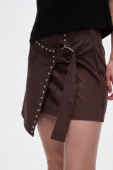 Minifalda marrón de gamuza sintética con corte irregular, detalle de tachas metálicas y cinturón con hebilla redonda.