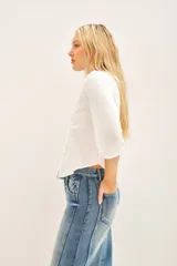 Camisa blanca de manga larga con cuello redondo y bordado floral en el delantero. El delantero es de algodón plano, mientras que las mangas y la espalda son de jersey a tono. Presenta un efecto fruncido en el centro de la espalda y puños en las mangas largas.