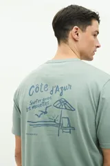 Remera verde de manga corta con cuello redondo y estampado en la espalda con la frase "Côte d'Azur Surfer avec les mouettes" y un dibujo de una sombrilla, una reposera y gaviotas.