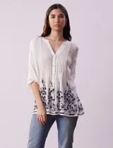 Blusa blanca de gasa bambula con cuello a la base, escote en V y pliegues. Tiene apertura central con botones, mangas ¾ con terminación fruncida y ruedo con detalles bordados en negro.