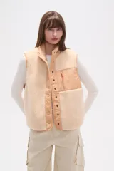 Chaleco holgado confeccionado en poliéster y corderito en tonos beige y crema. Presenta cuello alto, cierre frontal con botones a presión y un bolsillo con cierre en el pecho. Combina texturas de corderito con paneles de tela lisa.