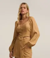 Blusa cropped color beige con escote corazón, mangas largas abullonadas con puños elásticos, botones frontales y detalle de volados en el ruedo.