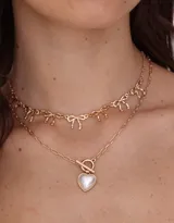 Collar corto doble, con cadena dorada de eslabones y cadena con dijes de moños. Incluye cierre marinero con dije de corazón de perla.