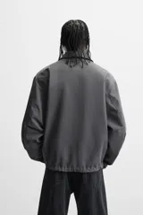 Cazadora relaxed fit color gris oscuro, confeccionada en tejido técnico repelente al agua. Tiene cuello solapa de cuero negro, manga larga con puños ajustables, bolsillos de vivo en la cadera, bolsillo interior, bajo ajustable con cordón y cierre frontal con cremallera oculta por solapa con botones.