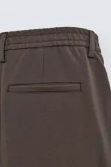 Pantalón negro de corte ancho con cintura elástica, pliegues frontales, bolsillos laterales y traseros de vivo. Tiene detalle de hebilla metálica en la trabilla y cierre frontal con cremallera y botón.