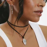 Collar con dije plateado en forma de máscara tribal, colgando de un cordón negro.