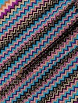 Poncho de tejido de punto con capucha y cierre frontal. Presenta el icónico patrón de zigzag (chevron) en múltiples colores vibrantes como azul turquesa, naranja, rosa, negro y beige. El borde inferior tiene flecos.
