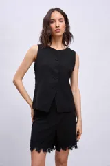 Chaleco negro sin mangas, con cuello redondo y cierre frontal de botones. Presenta detalles de encaje o apliques en los laterales y un corte ligeramente holgado.