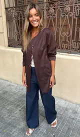 Chaqueta tipo blazer sin cuello, color marrón oscuro, de corte recto y largo a la cadera, con mangas tres cuartos y bolsillos frontales.