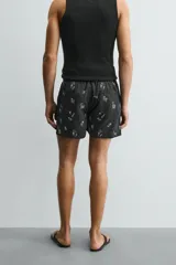 Bañador negro con estampado de dibujos en blanco. Tiene cintura elástica ajustable con cordón, bolsillos laterales y trasero.