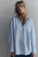 Camisa celeste de popelina de algodón, con cuello solapa, escote pico, manga larga con puños y cierre frontal con botones.