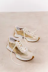 Zapatillas deportivas color beige con detalles metalizados dorados, confeccionadas en cuero con perforaciones, cordones ajustables y suela de goma dentada.