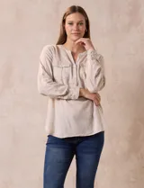 Blusa color beige de lino y rayón, con cuello a la base, escote en V y abertura con botones. Tiene bolsillos en la parte delantera y mangas largas regulables con presilla y botón.