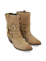 Bota corta de cuero gamuza color beige, con taco de madera y punta fina. Presenta tres tiras con hebillas metálicas plateadas y tachas pequeñas alrededor del tobillo.