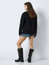 Chaqueta de jean negra de corte oversize, con cuello clásico, cierre frontal con botones metálicos, bolsillos delanteros con solapa y botón, y mangas largas.