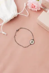 Pulsera de acero quirúrgico plateado con cadena de eslabones y dije circular blanco con borde negro y la letra "D" en negro.