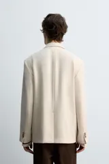 Blazer color beige confeccionado en tejido con lana, de corte relajado. Tiene cuello con solapas de muesca, manga larga con puños abotonados, bolsillo de vivo en el pecho, bolsillos de solapa en la cadera, bolsillo interior, bajo con abertura en el centro de la espalda y cierre frontal con botones.