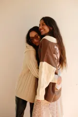 Buzo de punto en color marrón con cuello polo blanco y mangas largas blancas con textura de ochos. Presenta un diseño gráfico de gran tamaño en la espalda en tonos rosado, blanco y negro.