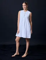 Vestido corto blanco de algodón y rayón, sin mangas, con cuello a la base, abertura central con botones y detalle plisado en la delantera. Incluye bolsillos laterales.