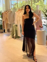 Vestido negro con escote strapless, corte ajustado al cuerpo y falda midi con abertura lateral y sobrefalda de tul transparente.
