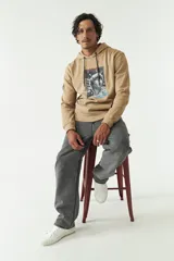 Canguro color beige con capucha y cordones ajustables, con bolsillo tipo canguro y puños y cintura acanalados. Presenta un estampado rectangular con una imagen y texto en el frente.