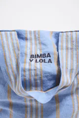 Bolso tipo shopper de lona color crudo con rayas verticales amarillas y grises. Tiene doble asa de hombro y logo de la marca estampado en el frente.