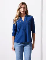 Blusa blanca de poliéster y elastano, con cuello a la base, escote en V y mangas tres cuartos. Presenta dos bolsillos frontales con cierre metálico dorado.