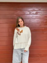 Sweater color crema tejido en lana merino, con apliques de cuero marrón y tejido trenzado en el frente. Presenta costuras a la vista en el ruedo y los puños.