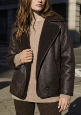 Chaqueta tipo gamulán de calce oversize en color marrón oscuro, con exterior de poliéster y forro de corderito sintético. Presenta cuello de solapa ancha y cierre frontal con botones.