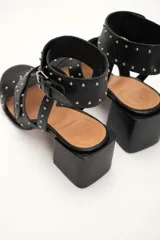 Sandalia de cuero vacuno color negro con tachas plateadas, tira ancha sobre los dedos, pulsera al tobillo con hebilla y taco cuadrado.