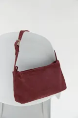Cartera de hombro color bordó, confeccionada en gamuza, con correa ajustable y hebilla metálica.