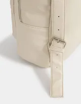Mochila de cuerina color crudo con bolsillo frontal con cierre y charm decorativo con cierre. Cuenta con correas regulables y manija superior.