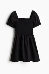 Vestido corto negro de popelina de algodón con escote cuadrado y mangas cortas abullonadas con elástico. El cuerpo es ajustado con fruncido y la falda con vuelo. Tiene ribete de volado en escote y puños.