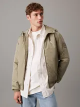 Campera Calvin Klein color verde oliva, con capucha, cierre frontal con cremallera, puños y bajo elásticos, bolsillos con botón de presión en la cintura y logo de la marca en el pecho y la manga. Confeccionada en nailon 100% reciclado y forro de poliéster.