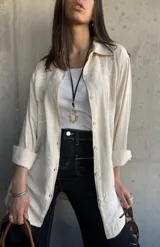Camisa beige con bordados florales blancos, de corte recto y mangas largas con puños enrollados.