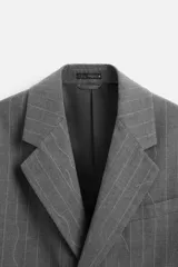 Blazer gris con raya diplomática, corte boxy, cuello con solapas de muesca, manga larga con puño abotonado, bolsillo de vivo en pecho, aberturas en espalda y cierre frontal con dos botones.