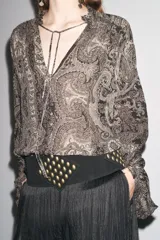 Camisa de viscosa con estampado paisley en blanco y negro. Cuello redondo con escote pico ajustable con cordones, mangas largas con elástico y volantes en los puños, bajo asimétrico y cierre frontal con botones forrados.