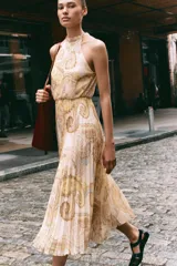 Vestido midi de cuello halter con estampado paisley en tonos beige, amarillo y marrón. Tiene lazada en la espalda, cintura elástica y falda plisada. Cierre trasero con cremallera oculta, abertura y botón.