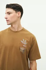 Remera color marrón con cuello redondo y manga corta. Presenta un bordado en el lado izquierdo del pecho con un diseño abstracto y texto.
